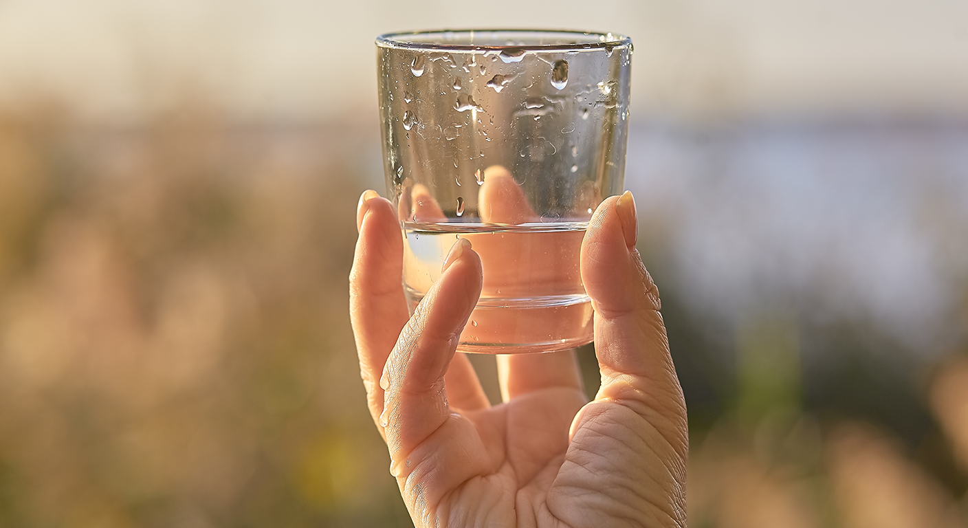 A hand holding a glass of water outdoors in warm sunlight